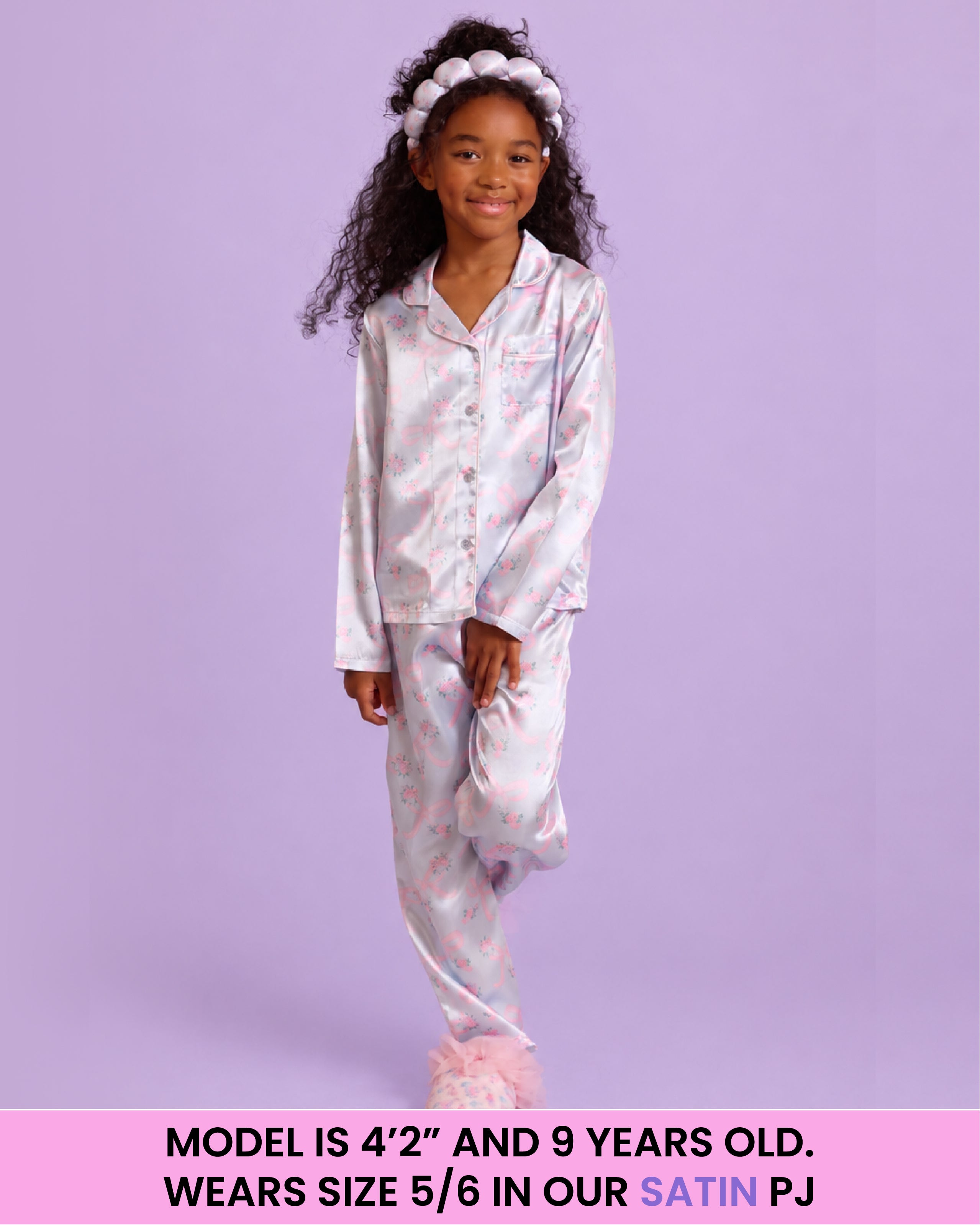 A young girl models a two-piece satin pajama set featuring a light blue base with a pink floral pattern, set against a solid lavender background. A pink banner at the bottom contains sizing information.