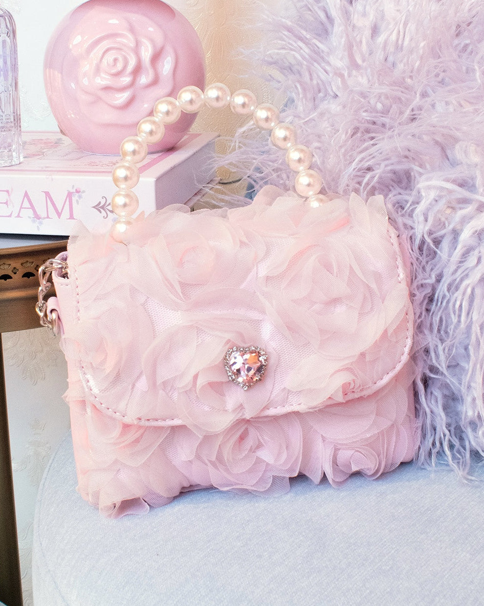 Pink handbag with floral details and a decorative brooch on a light blue surface, with a pink lamp and feathered decoration in the background.