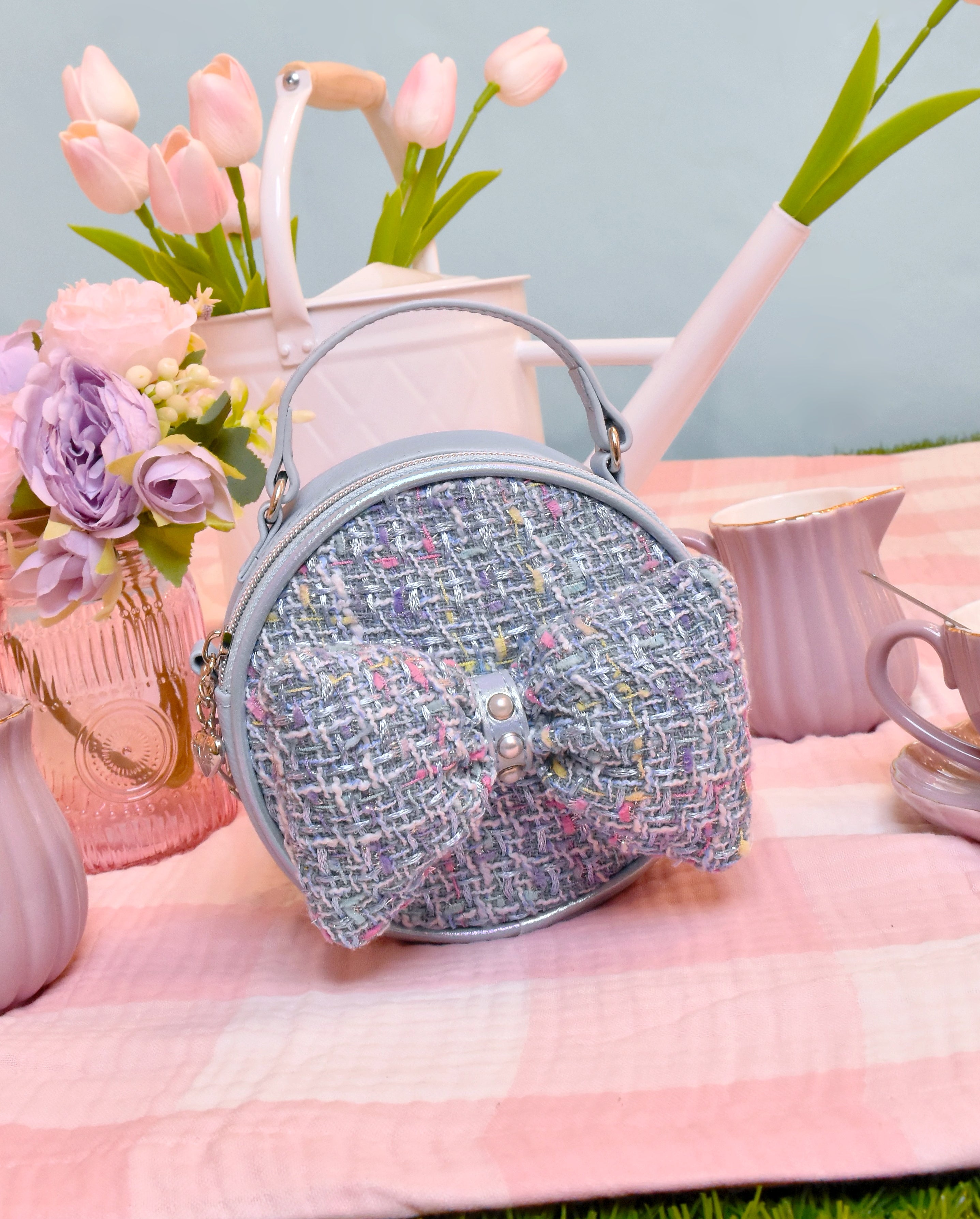 front facing view of a circular blue tweed mini bag with a top handle and matching blue tweed bow detail on the front placed on top of a pink and white checkered picnic blanket