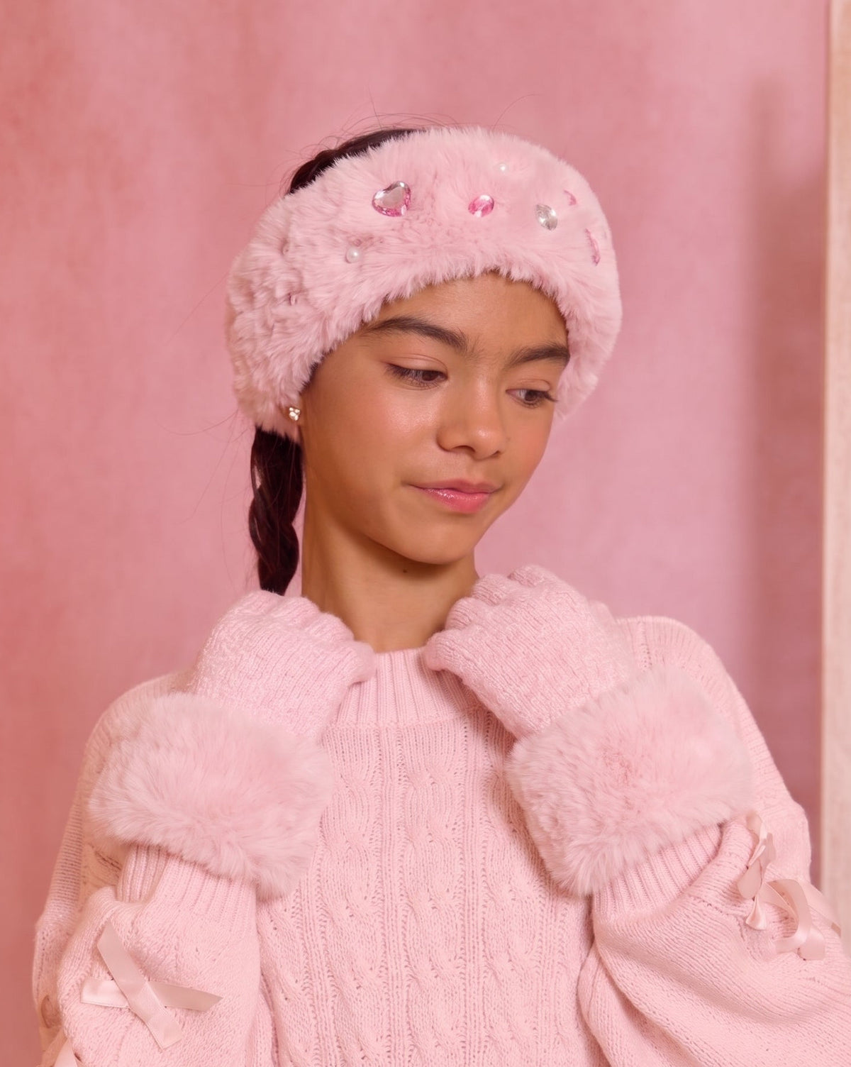 A girl wearing a pink knit and faux-fur accessory set including a rectangular headband with rhinestone embellishments and a pair of knit gloves with fluffy trim.