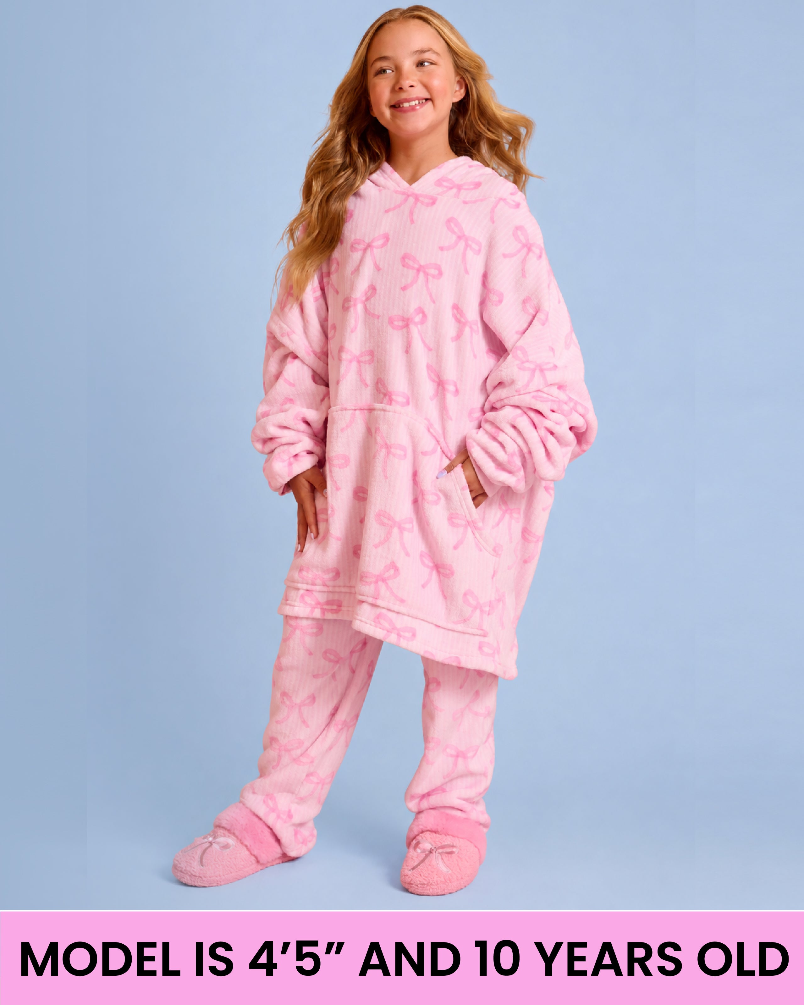 A young girl wearing a pink hooded pajama set with an allover bow pattern, standing against a light blue background. A pink banner at the bottom reads 'MODEL IS 4'5" AND 10 YEARS OLD'.