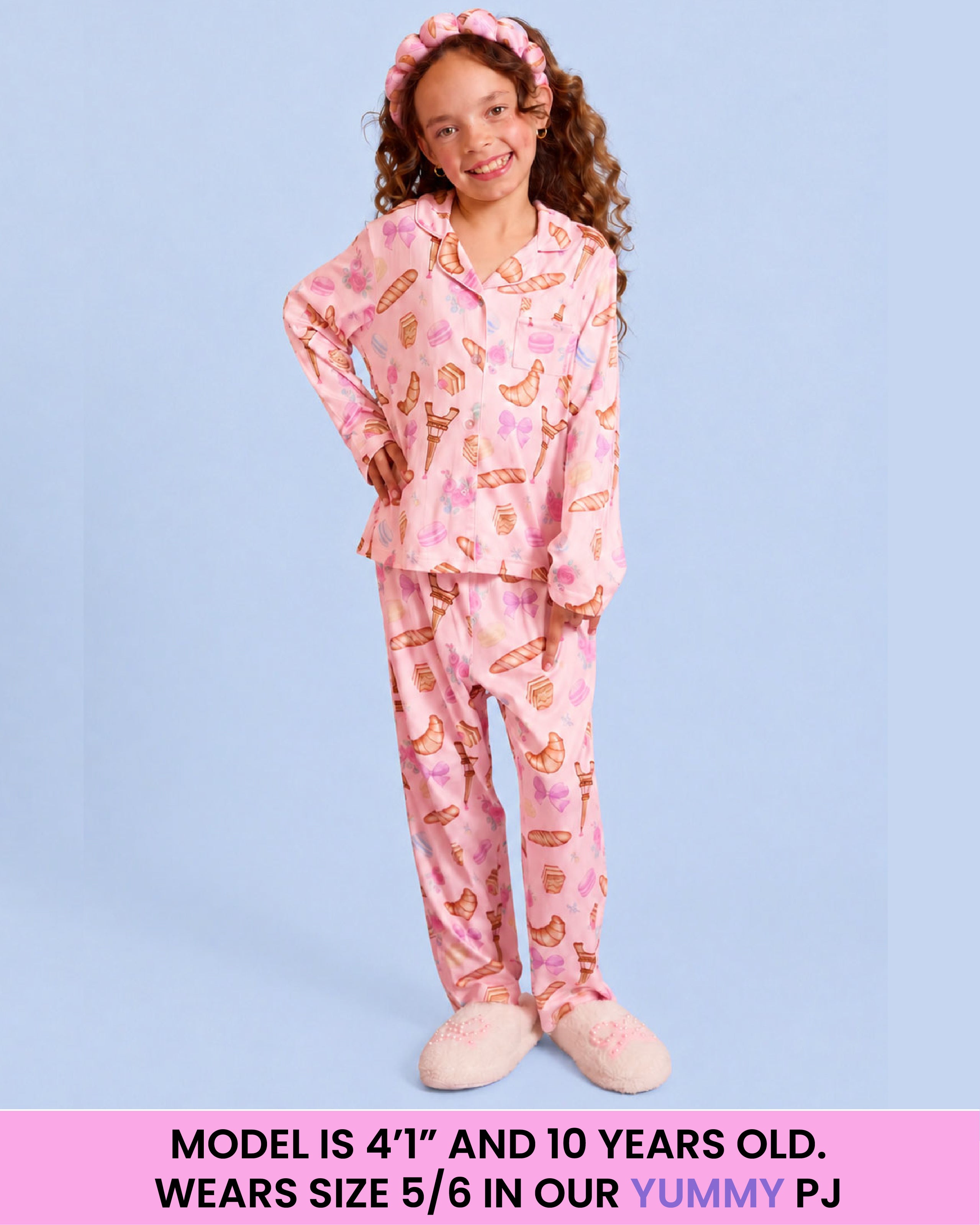 A young girl modeling a pink pajama set with a Paris-themed print, consisting of a long-sleeve top and matching pants, paired with a matching pink headband and slippers. The image has a text banner at the bottom with sizing information.