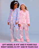 Two girls modeling matching pajama sets against a light blue-gray studio background. The left model wears a light blue robe with pink bow patterns and pink slippers, and the right model wears a pink robe with pink bow patterns and pink slippers.