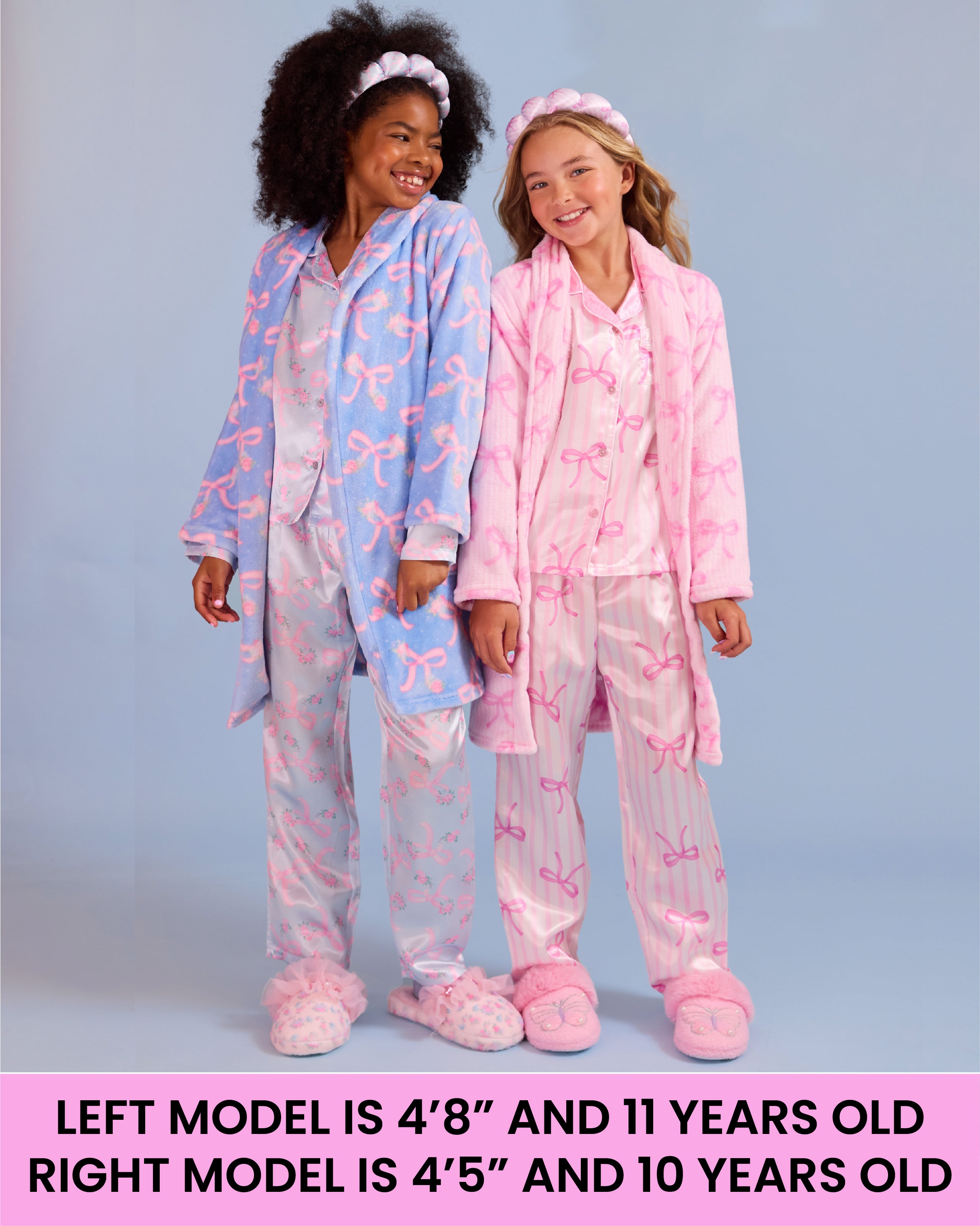Two girls modeling matching pajama sets against a light blue-gray studio background. The left model wears a light blue robe with pink bow patterns and pink slippers, and the right model wears a pink robe with pink bow patterns and pink slippers.