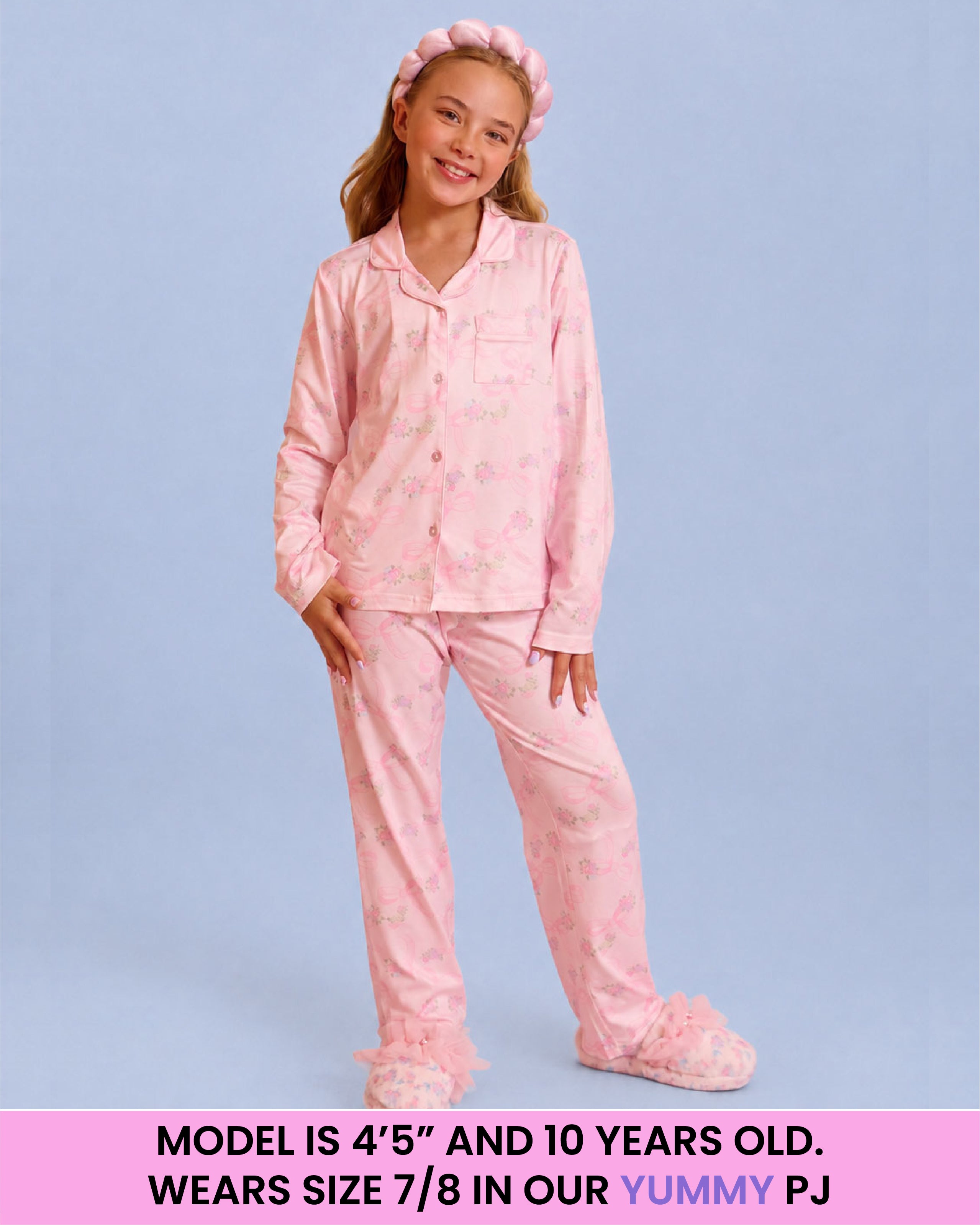 A young girl modeling a light pink pajama set with a subtle floral pattern, matching pink headband, and pink slippers against a solid light blue background. A pink banner at the bottom contains sizing information.