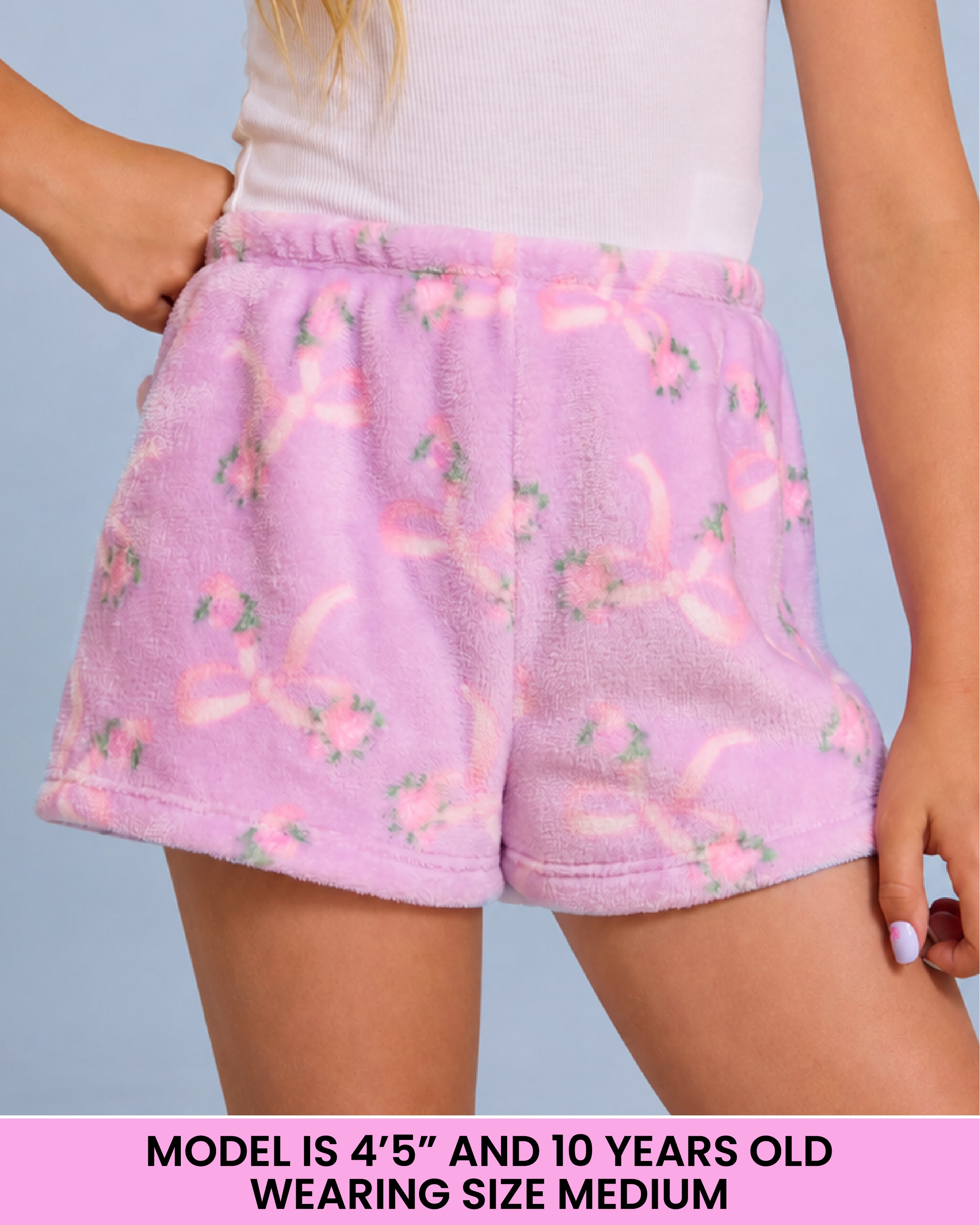 A person wearing light purple plush shorts with a pink ribbon and floral pattern, shown from the waist to mid-thigh against a pale blue background. A text banner at the bottom reads, 'MODEL IS 4'5" AND 10 YEARS OLD WEARING SIZE MEDIUM'.
