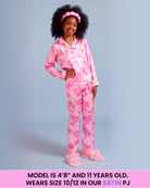 A young girl with dark curly hair wearing a pink floral satin pajama set, consisting of a long-sleeve button-up top and matching pants, standing against a light blue background. A pink text banner is at the bottom with sizing information.