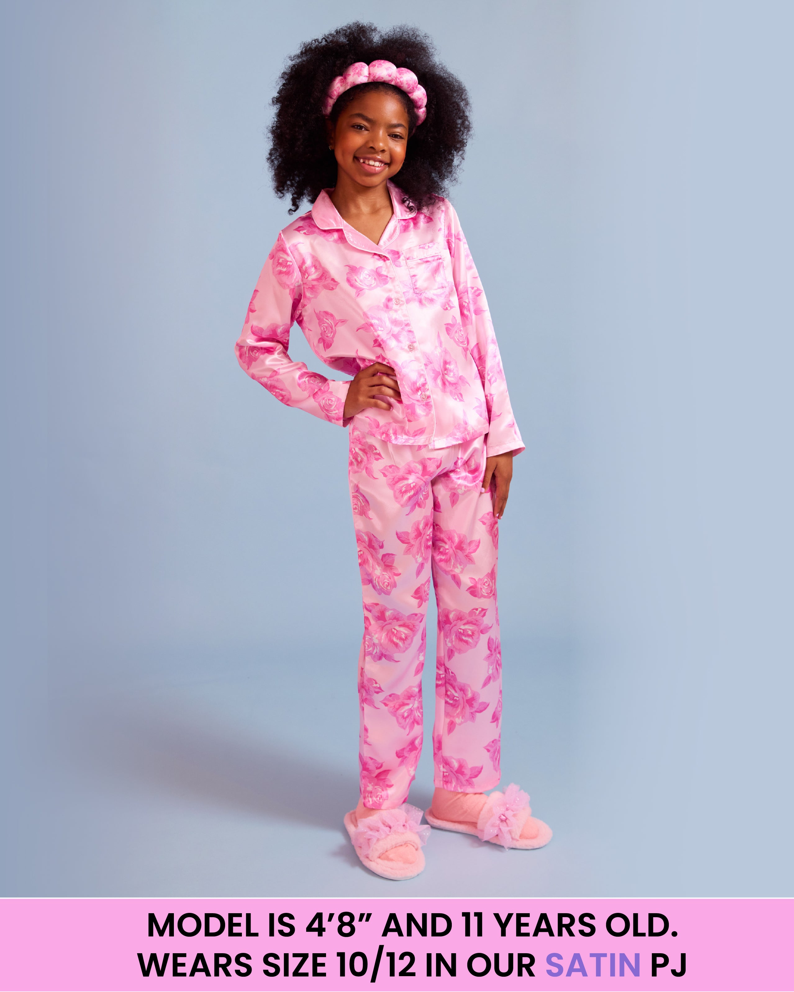 A young girl with dark curly hair wearing a pink floral satin pajama set, consisting of a long-sleeve button-up top and matching pants, standing against a light blue background. A pink text banner is at the bottom with sizing information.