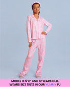 A young girl modeling a purple and white checkered pajama set consisting of a long-sleeve top and matching pants, with a matching pink bow headband and fluffy slippers, set against a solid light blue background with a text banner at the bottom.