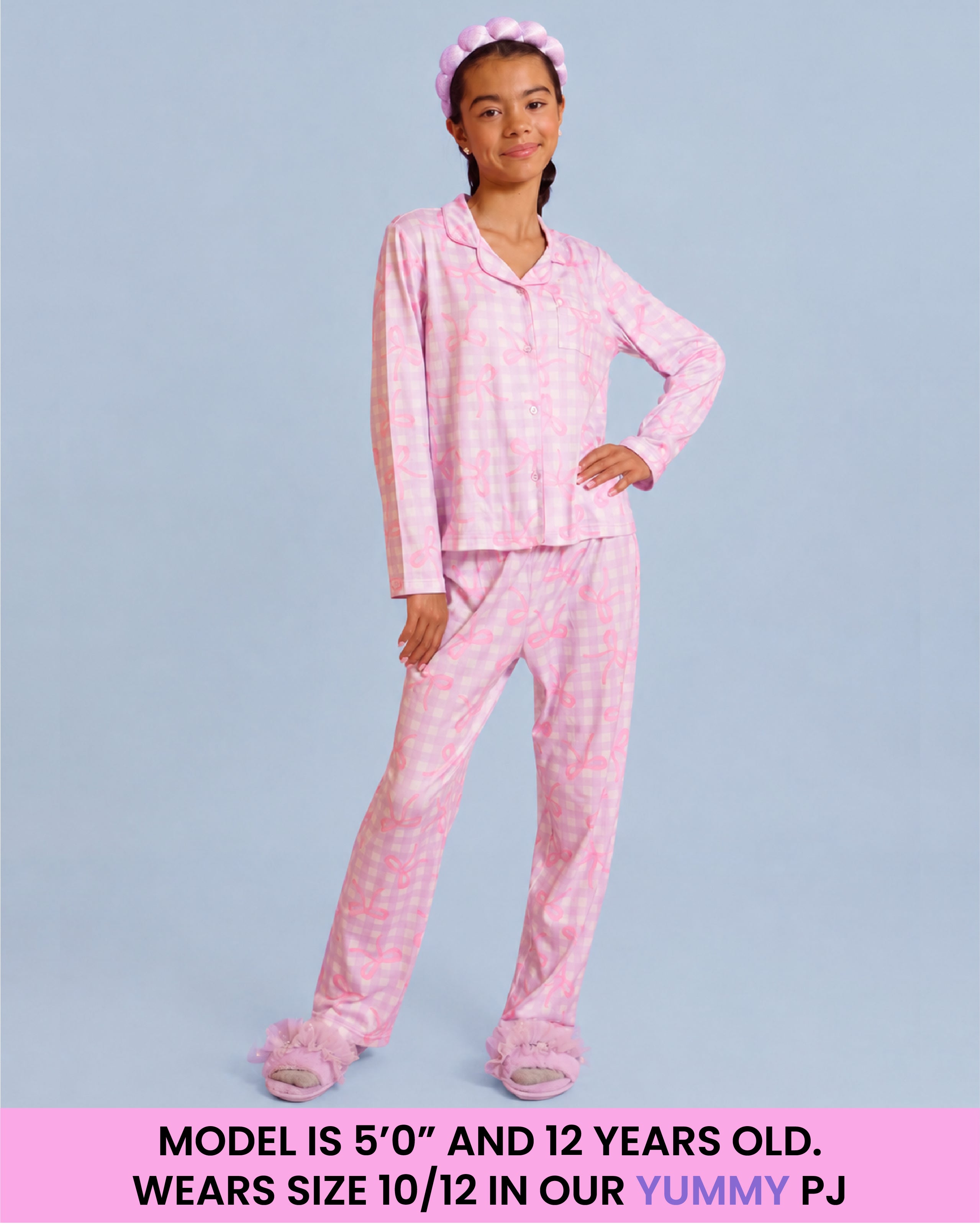 A young girl modeling a purple and white checkered pajama set consisting of a long-sleeve top and matching pants, with a matching pink bow headband and fluffy slippers, set against a solid light blue background with a text banner at the bottom.