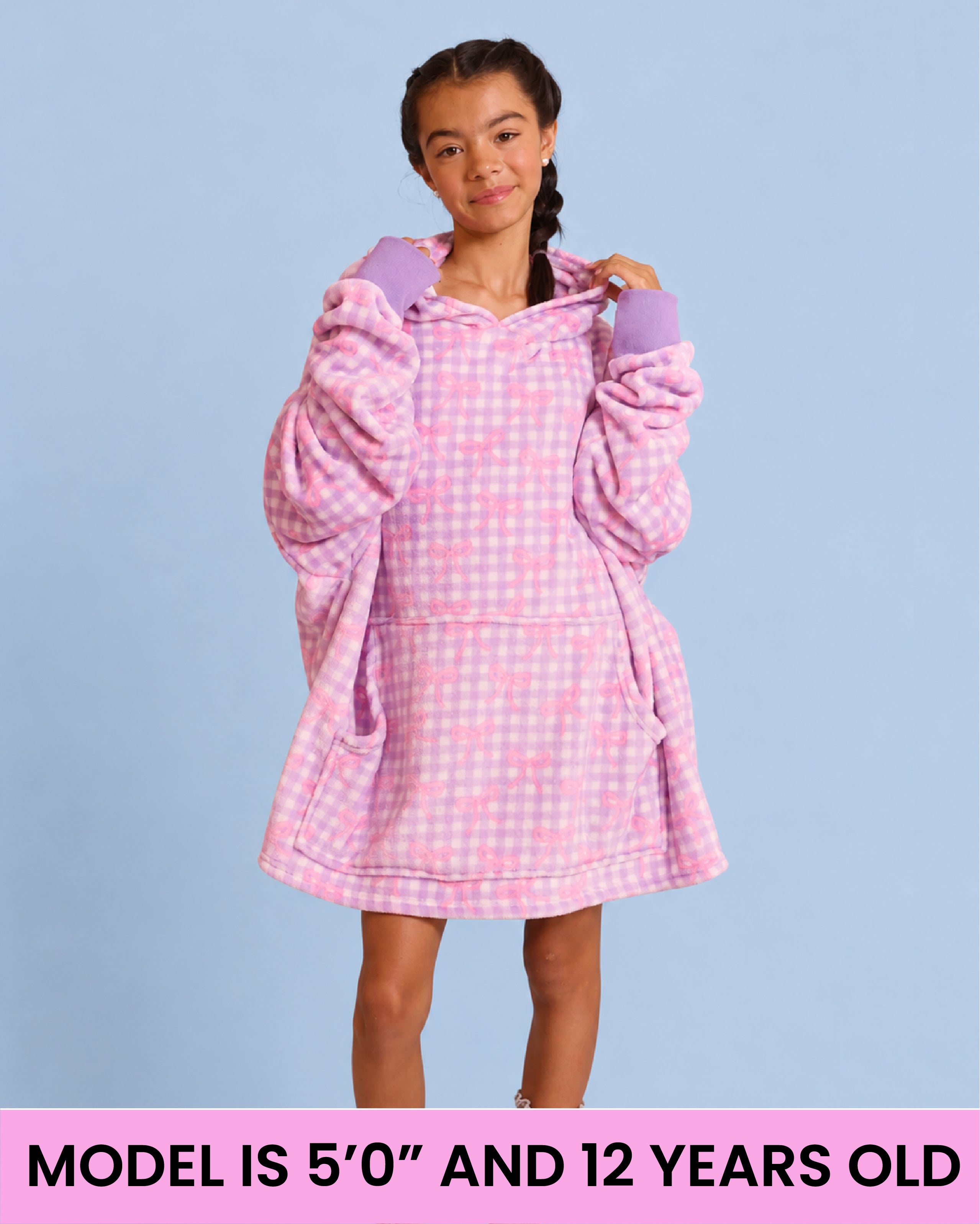 A young girl wearing a purple and white gingham plush hooded robe with long sleeves, standing against a light blue background. A pink banner at the bottom reads 'MODEL IS 5'0" AND 12 YEARS OLD'.