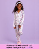 A young girl models a two-piece satin pajama set featuring a light blue base with a pink floral pattern, set against a solid lavender background. A pink banner at the bottom contains sizing information.