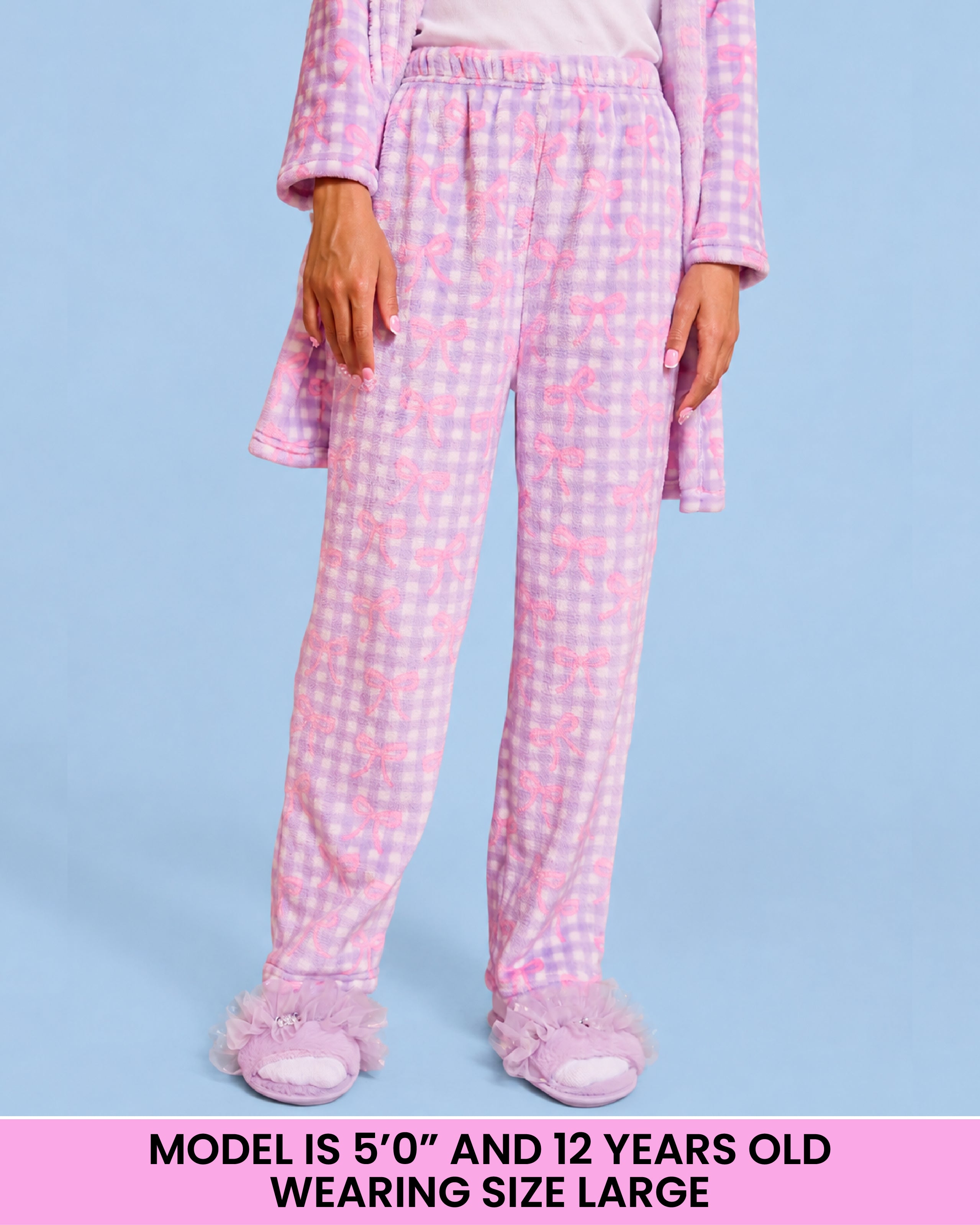 A child wearing purpleand white gingham pajama pants with a bow pattern, shown from the waist down against a light blue background. A pink text banner at the bottom reads 'MODEL IS 5'0" AND 12 YEARS OLD WEARING SIZE LARGE'.