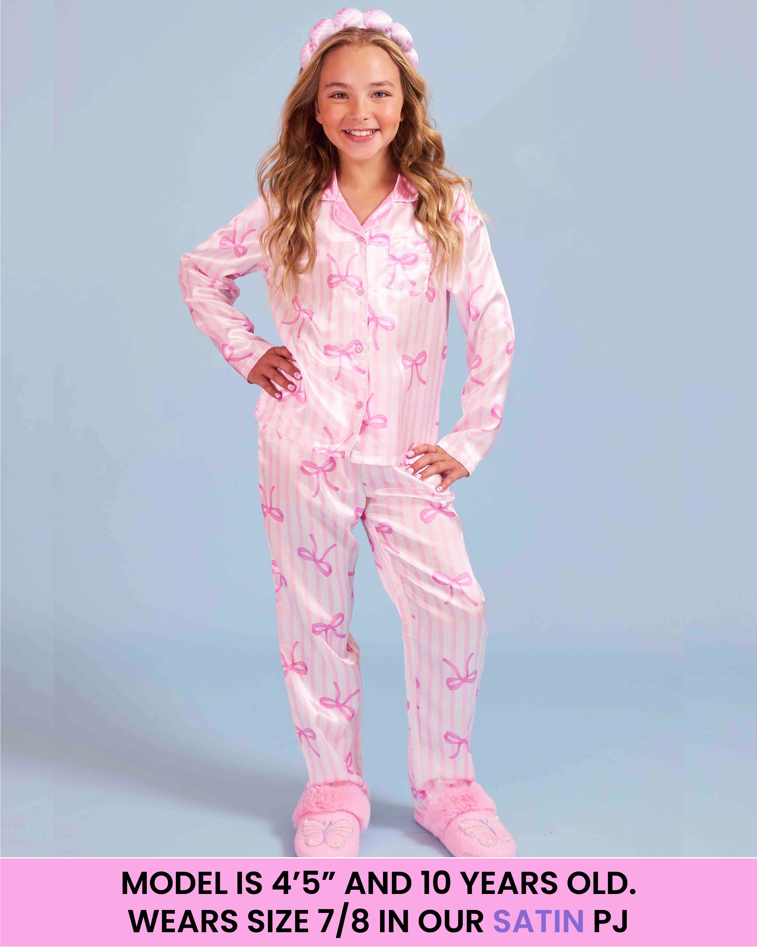 A young girl modeling a pink pajama set with a striped bow pattern, consisting of a long-sleeve top and matching pants, along with a matching headband and slippers. The image has a text overlay at the bottom describing the model's age and size.