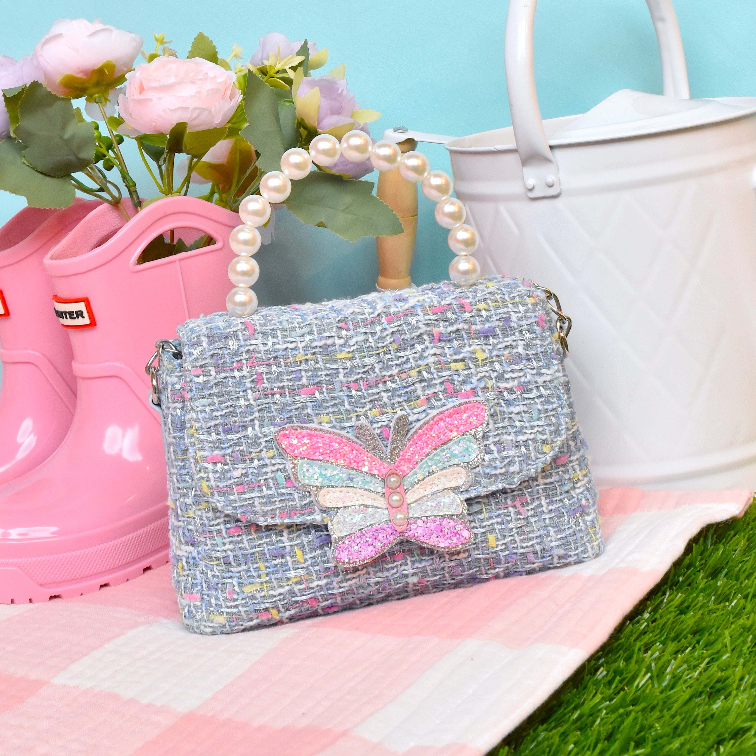 front facing view of a blue tweed mini bag with a pearl handle and glitter butterfly appliqué on the front flap sitting on top of a picnic blanket