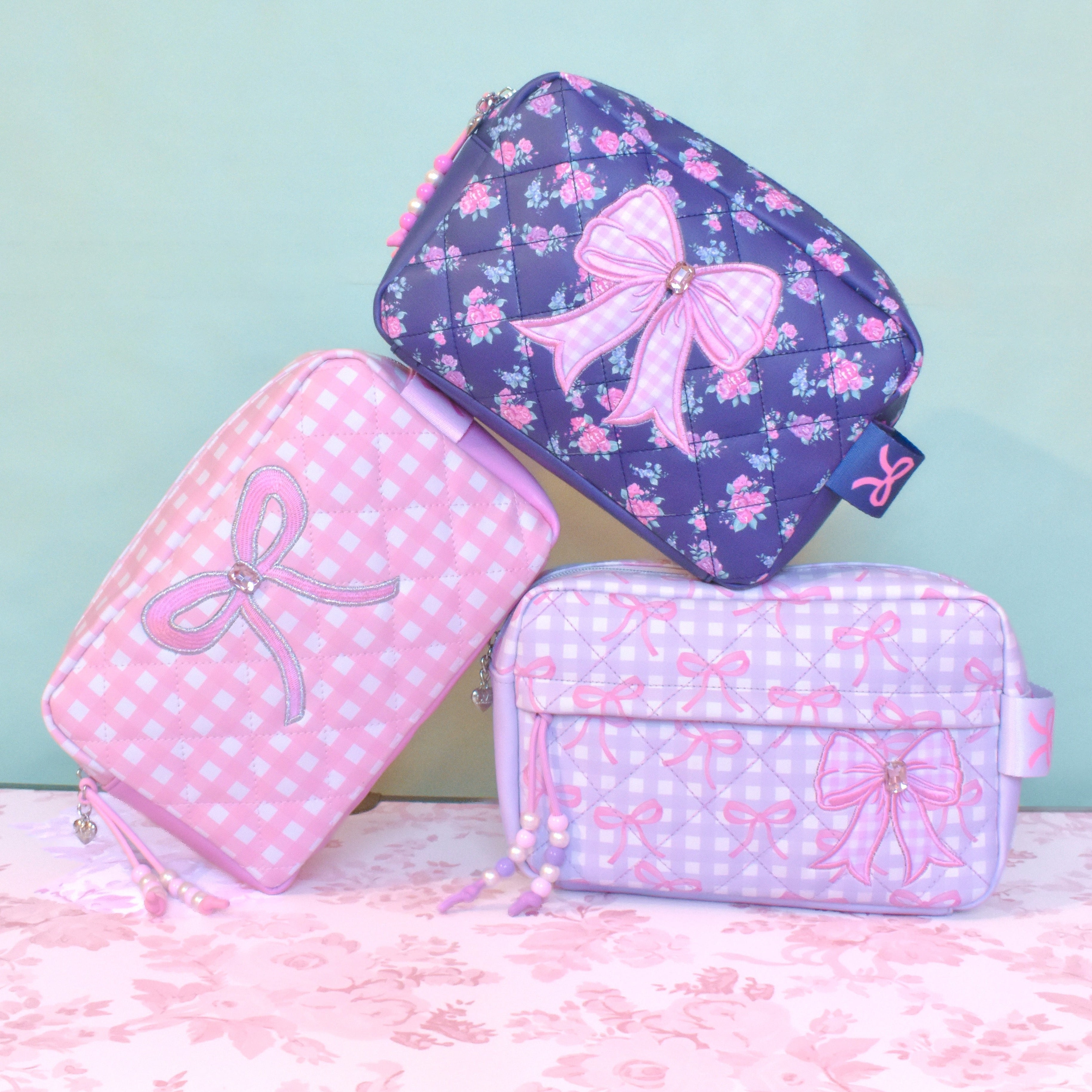 Three pink and purple cosmetic bags with bow designs on a light green background.