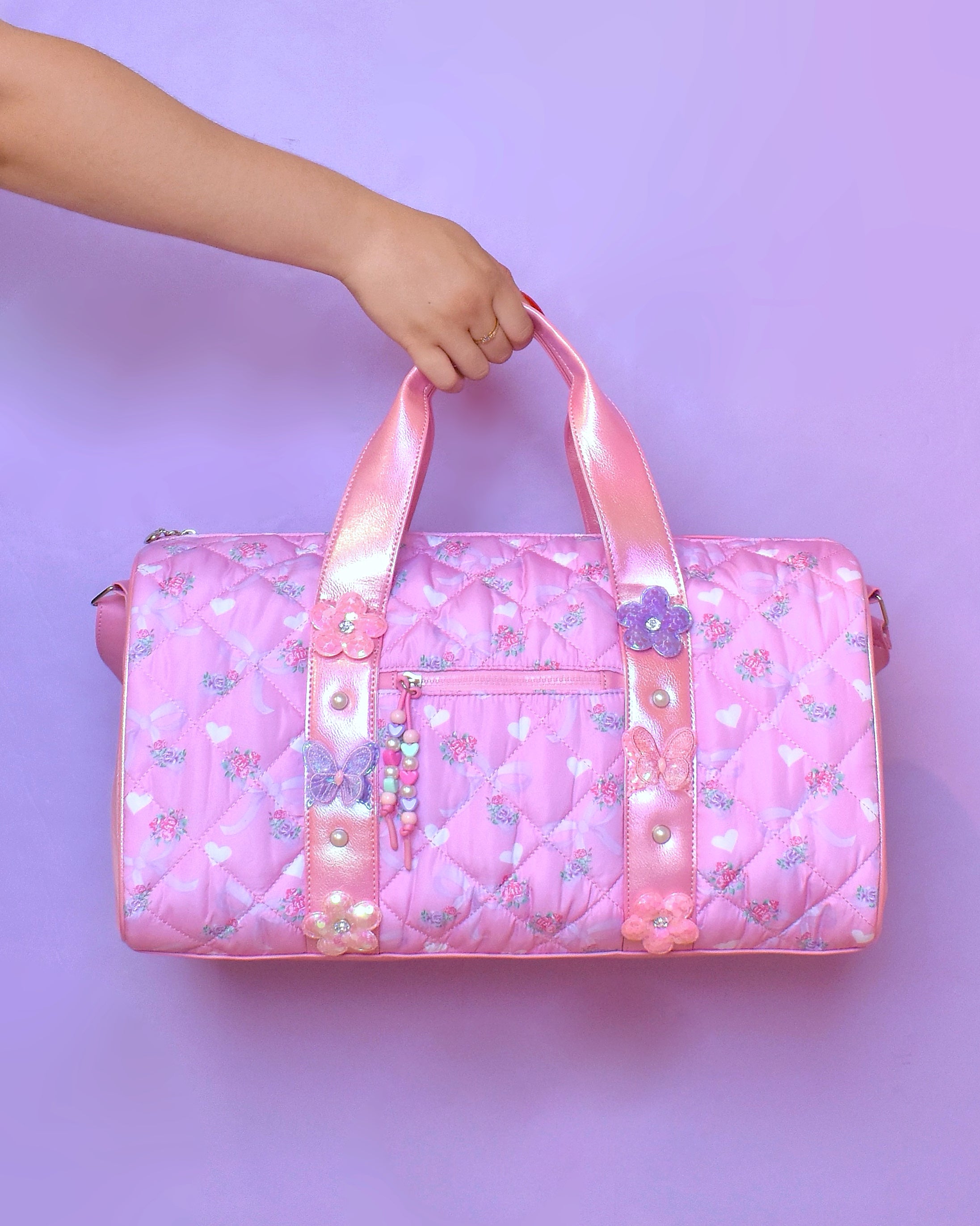 front view of a floral bow printed barrel duffle bag with butterfly & daisy appliques. pink metallic straps & beaded zipper pull