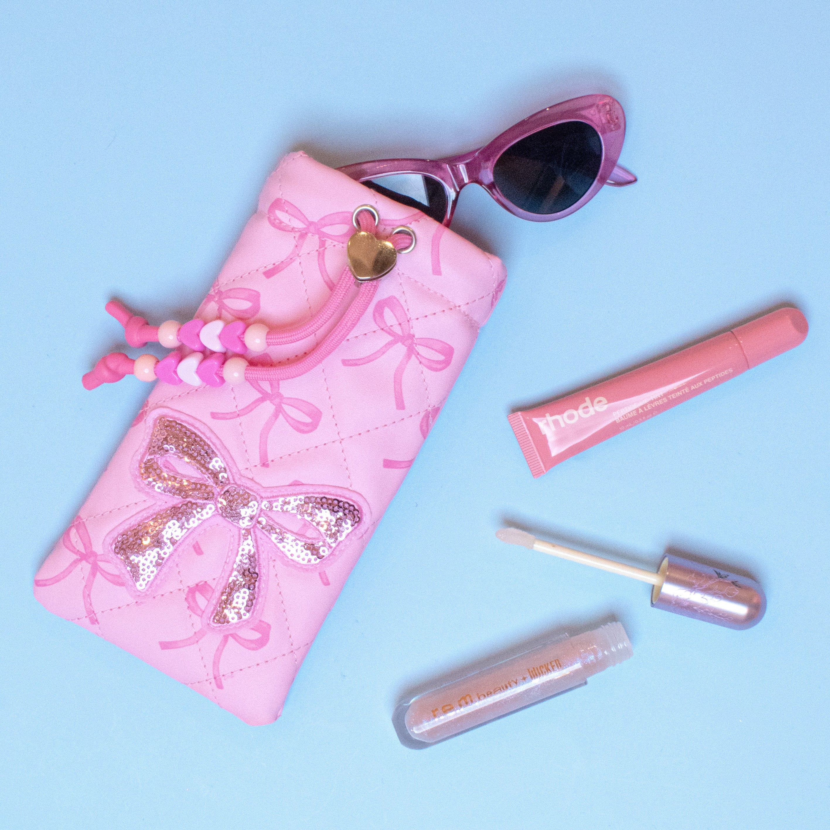 Pink sunglass pouch with aglittery bow, sunglasses, and lip gloss on a light blue background