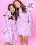 Two girls wearing matching purple floral bow print oversized blanket hoodies with a pink background and 'Holiday Best Seller' label.
