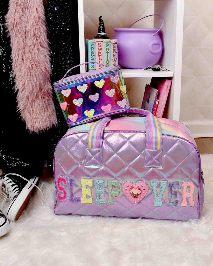 Lifestyle image of purple quilted metallic large 'Sleepover' duffle sitting in front of girl's fall-themed wardrobe
