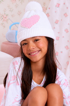 A smiling young girl wearing a white knit pom-pom beanie with a pink heart on the front, paired with a matching white and pink patterned shirt, sitting in a cozy room with floral wallpaper.