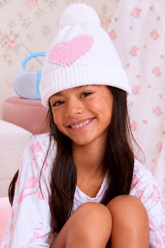 A smiling young girl wearing a white knit pom-pom beanie with a pink heart on the front, paired with a matching white and pink patterned shirt, sitting in a cozy room with floral wallpaper.
