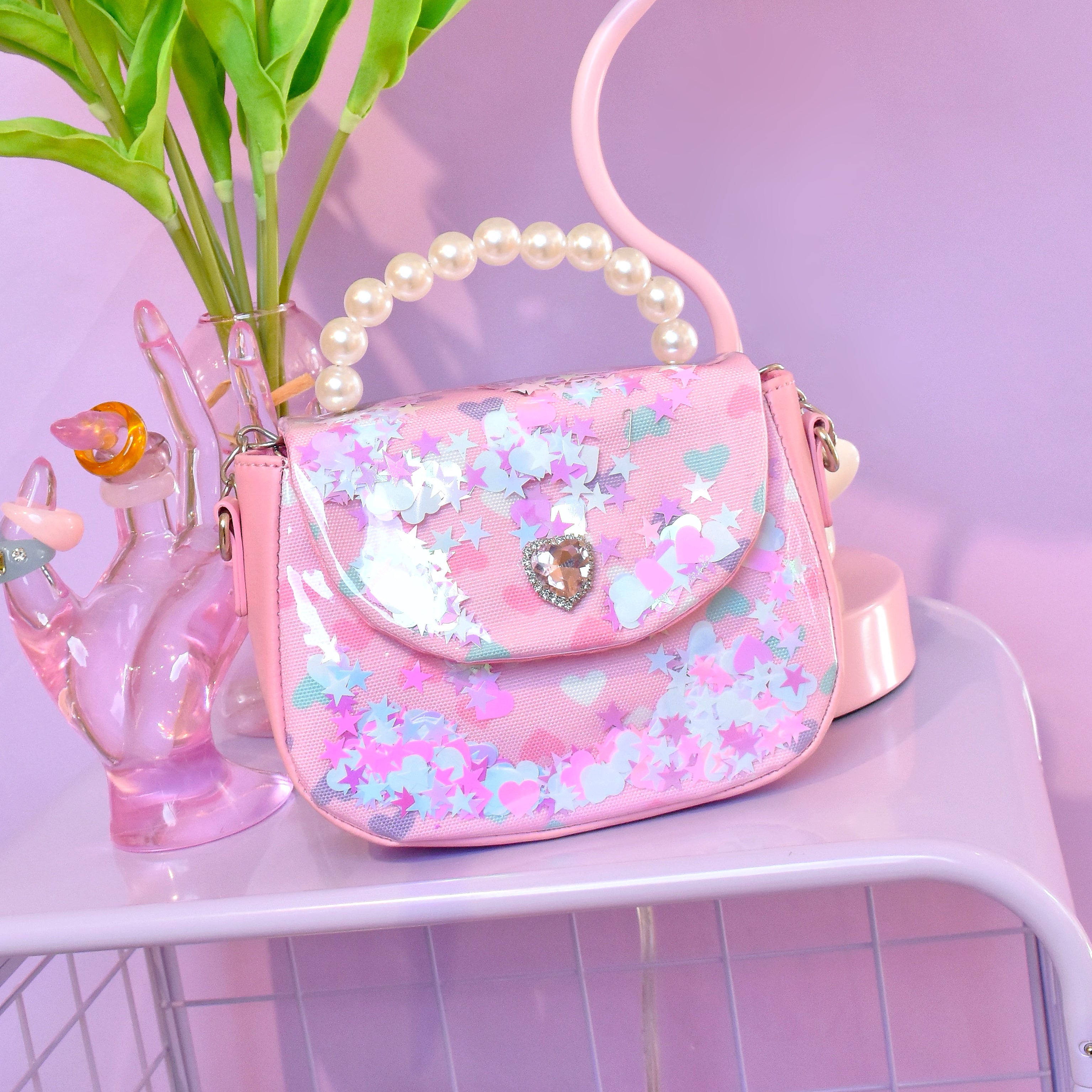 front facing view of a vinyl mini bag with a a flap front opening, pearl top handle, printed hearts pattern, and loose confetti detailing. the bag is sitting on top of a purple metal shelf.