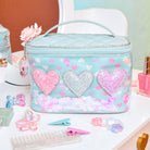 front facing view of a light blue vinyl round train case bag with top handle and heart charm placed on top of a vanity. the bag is detailed with printed hearts, loose confetti, and glitter heart appliqués, and a pink crystal heart charm.
