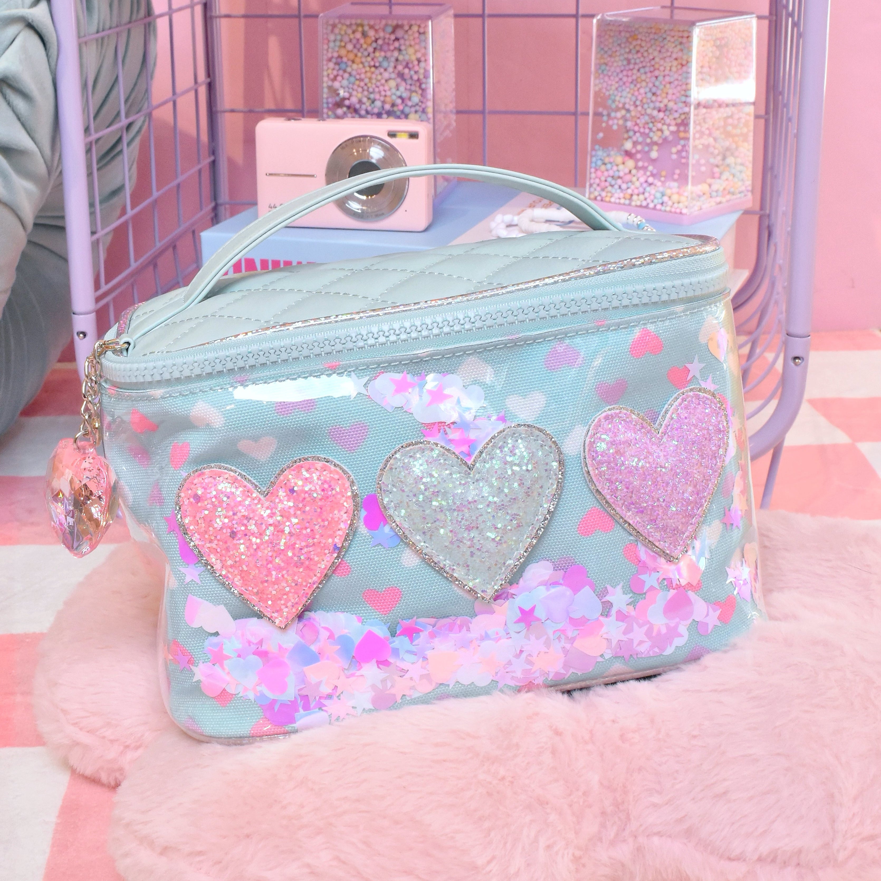 front facing view of a light blue vinyl round train case bag with top handle and heart charm placed on top of a pink flower shaped pillow. the bag is detailed with printed hearts, loose confetti, and glitter heart appliqués, and a pink crystal heart charm.