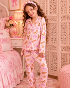 A young girl with long curly brown hair stands in a pink bedroom wearing a light pink pajama set with a pattern of macarons, bows, and hearts, along with a matching headband and fluffy slippers.