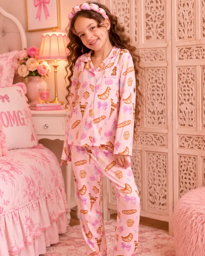A young girl with long curly brown hair stands in a pink bedroom wearing a light pink pajama set with a pattern of macarons, bows, and hearts, along with a matching headband and fluffy slippers.